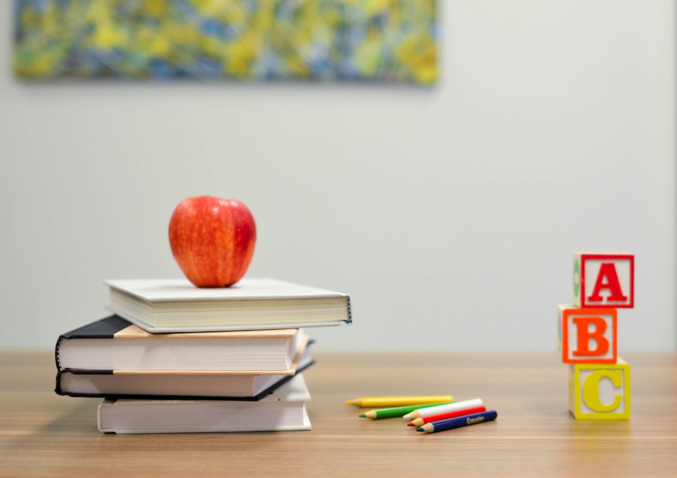 Resources 2 Stack of books with an apple, ABC blocks, and coloured pencils on a wooden desk in a classroom setting.